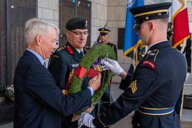 An elderly man in a dark blue suit and two military officers in dark blue dress uniforms hold a wreath between them as they place it on a war memorial.