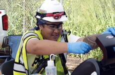 A woman in a yellow reflective vest, white hard hat and blue rubber gloves takes a water sample from a well.