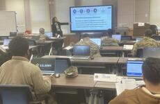 An instructor points at coursework on a large screen at the front of a room of military service members seated at tables with laptop computers.