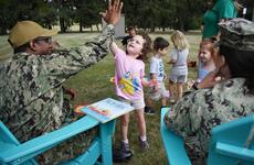 Two service members sit in chairs on a lawn with a group of children standing in front of them.