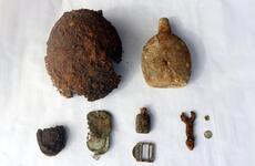 A collection of rusted and corroded wartime artifacts displayed on a white surface, including a damaged helmet, a canteen, small bottles, a buckle, a wrench and various metal fragments.