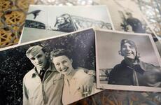 Black-and-white, 1940s-era photographs lie on a table depicting an aviator in a leather jacket and helmet and a man in a military dress uniform posing with his arm around a smiling woman in white.