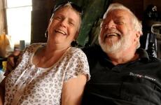 An elderly couple, seen from the waist up, laugh while seated next to each other.