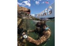 A diver in scuba gear is seen just below the surface of water next to the wreckage of a ship, with the sky above the waterline at the top of the scene.