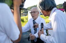 An elderly Japanese man wearing a white shirt, green tie and dark-rimmed glasses stands with the assistance on a cane, while another man to the right holds a microphone in front of him and a person to the left records with a video camera.