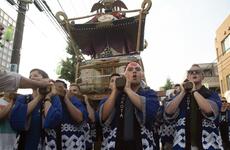A group of men in traditional blue and white patterned Japanese festival clothing (happi coats) carry a portable shrine (mikoshi) on their shoulders during a street festival. The scene takes place on a city street with buildings and power lines visible in the background.