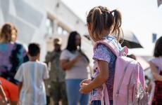 A girl wearing pigtails and wearing a pink backback looks around a school yard.