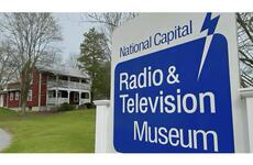 A closeup of a sign that reads “National Capital Radio & Television Museum” with a farmhouse in the background.