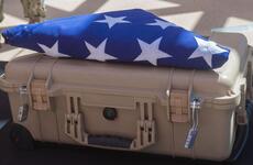A folded American flag with white stars visible on blue fabric rests on top of a tan military-style hard case. The flag is ceremonially folded in the traditional triangular manner typically used for military honors and funeral services. 