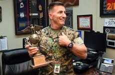 A Marine in camouflage uniform holds his left fist to his chest and holds a trophy in his right hand while standing in an office with framed memorabilia on the walls behind him.