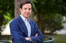 A man wearing a blue blazer and light-colored shirt stands outdoors with his arms crossed, looking at the camera. There are green trees and a blurred background behind him.