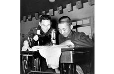 A black and white photo of a man looking on as a boy works a sewing machine on a table.
