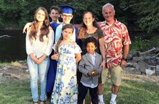 A family consisting of a father, mother and five children group together to pose on a grass lawn, with trees in the background.