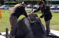 Two men in military dress uniforms place a yellow and black ribbon on a large rock with a memorial plaque on it.
