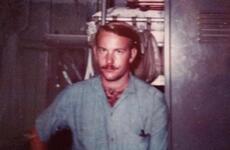 A veteran with a mustache and blue shirt stands in front of a brown background.