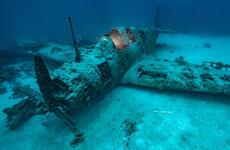 A wrecked, World War II-era military propeller plane sits on the ocean floor covered in sand and barnacles.