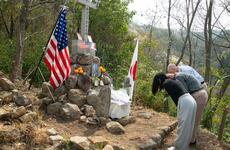 Three people bow in front of a hillside stone memorial displaying U.S. and Japanese flags, with offerings and flowers placed at its base.