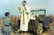 Kapaun stands above a soldier, with a jeep behind him.