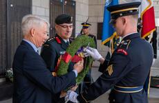 An elderly man in a dark blue suit and two military officers in dark blue dress uniforms hold a wreath between them as they place it on a war memorial.