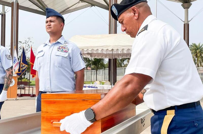 A soldier in dress uniform and white gloves places a wooden case into a casket, while another soldier in dress uniform stands at attention next to the casket.