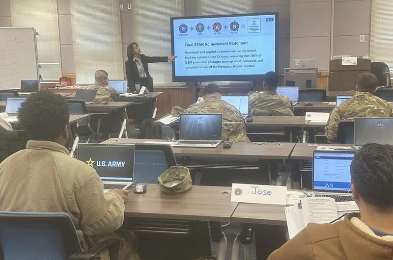 An instructor points at coursework on a large screen at the front of a room of military service members seated at tables with laptop computers.