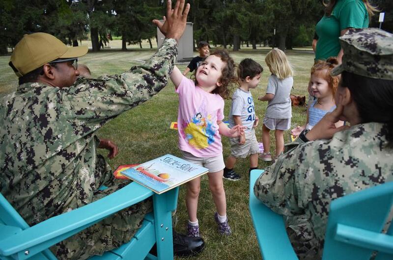 Two service members sit in chairs on a lawn with a group of children standing in front of them.