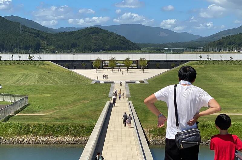 A Japanese man and boy, seen from behind, look out over a memorial park with a mountain scene in the background.