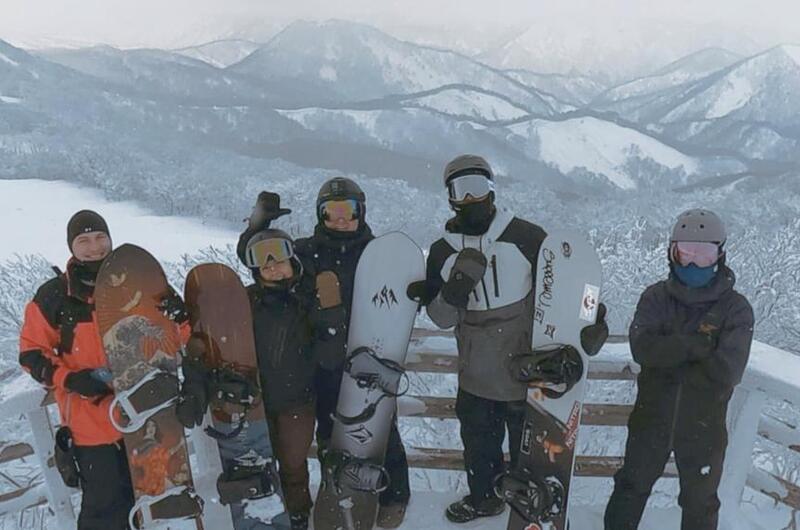 Five men holding snowboards pose for a picture in front of some mountains.