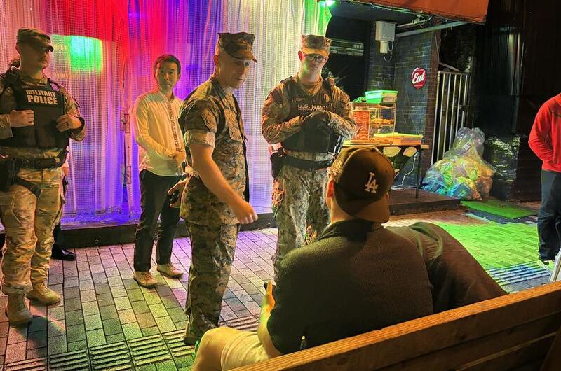 Military officials in camouflage uniforms speak to service members in civilian clothes seated on a street bench at night, near a colorful curtain backdrop.