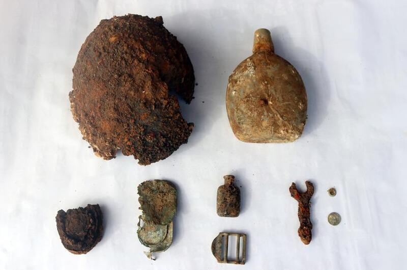 A collection of rusted and corroded wartime artifacts displayed on a white surface, including a damaged helmet, a canteen, small bottles, a buckle, a wrench and various metal fragments.