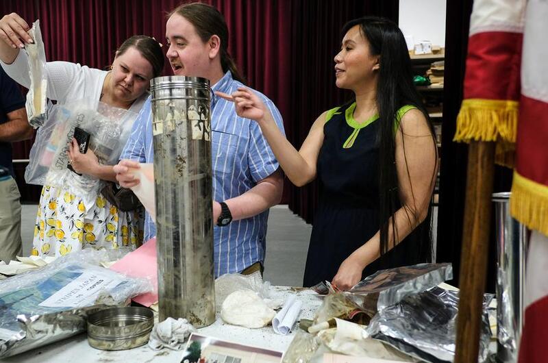 Three adults look at items in plastic bags that were put into a silver tube time capsule when they were children.
