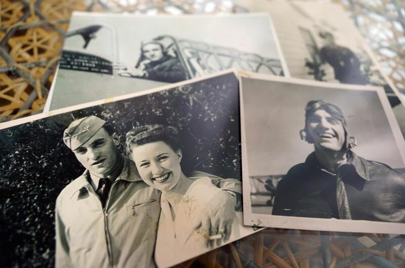 Black-and-white, 1940s-era photographs lie on a table depicting an aviator in a leather jacket and helmet and a man in a military dress uniform posing with his arm around a smiling woman in white.