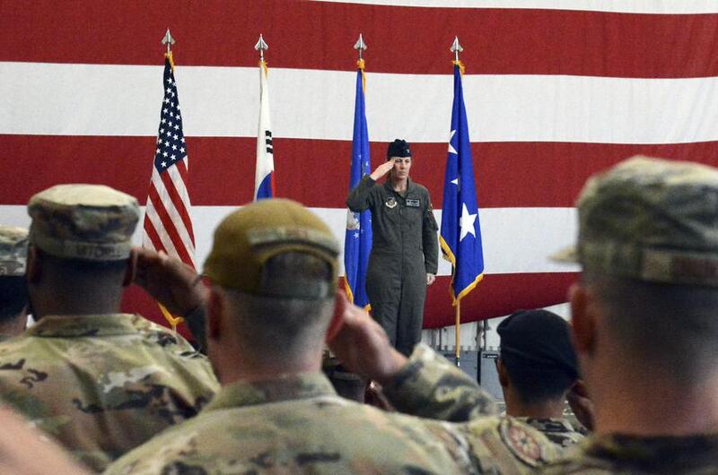 A military officer in flight suit salutes while standing at attention in front of four flags including the American flag and Air Force flags. The officer is positioned against a large American flag backdrop with red and white stripes. In the foreground, seated military personnel in camouflage uniforms and caps are saluting back.