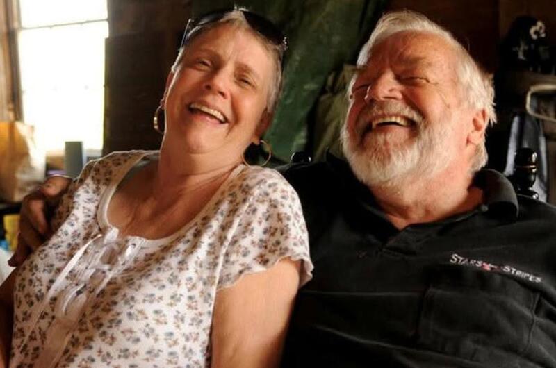 An elderly couple, seen from the waist up, laugh while seated next to each other.