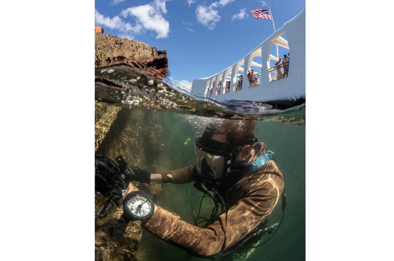 A diver in scuba gear is seen just below the surface of water next to the wreckage of a ship, with the sky above the waterline at the top of the scene.