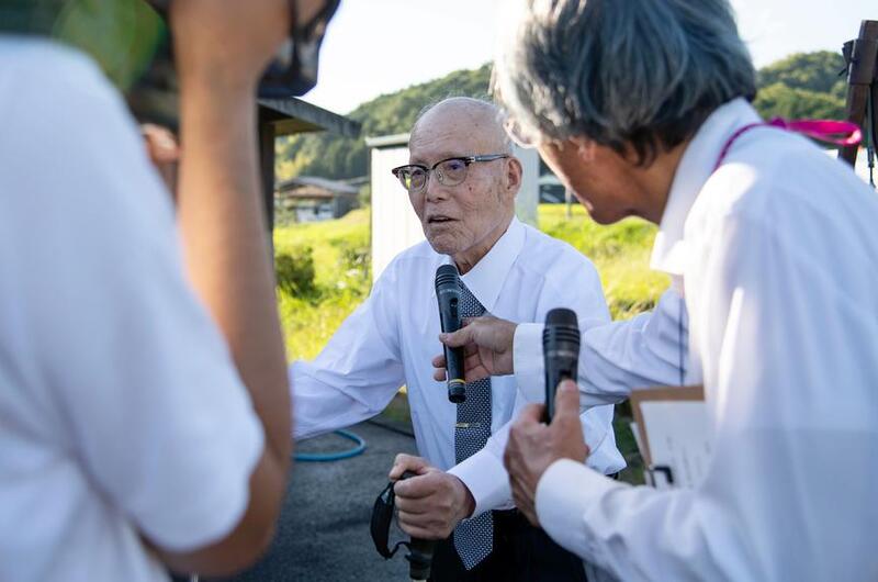 An elderly Japanese man wearing a white shirt, green tie and dark-rimmed glasses stands with the assistance on a cane, while another man to the right holds a microphone in front of him and a person to the left records with a video camera.