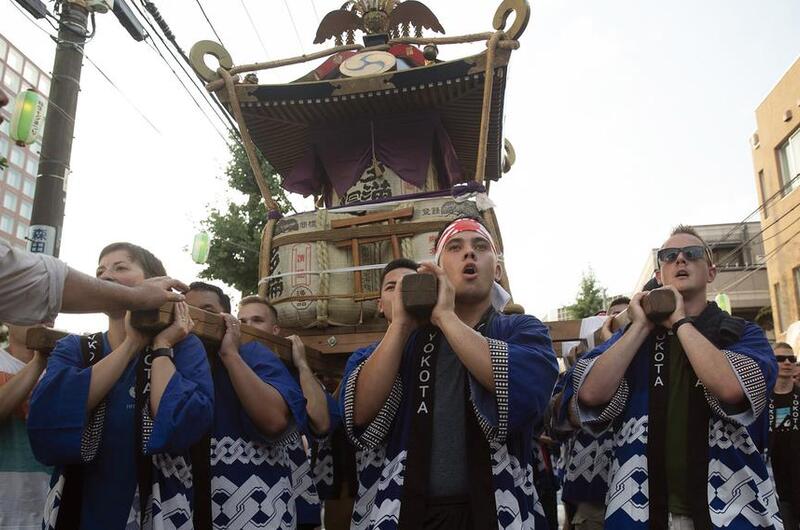 A group of men in traditional blue and white patterned Japanese festival clothing (happi coats) carry a portable shrine (mikoshi) on their shoulders during a street festival. The scene takes place on a city street with buildings and power lines visible in the background.