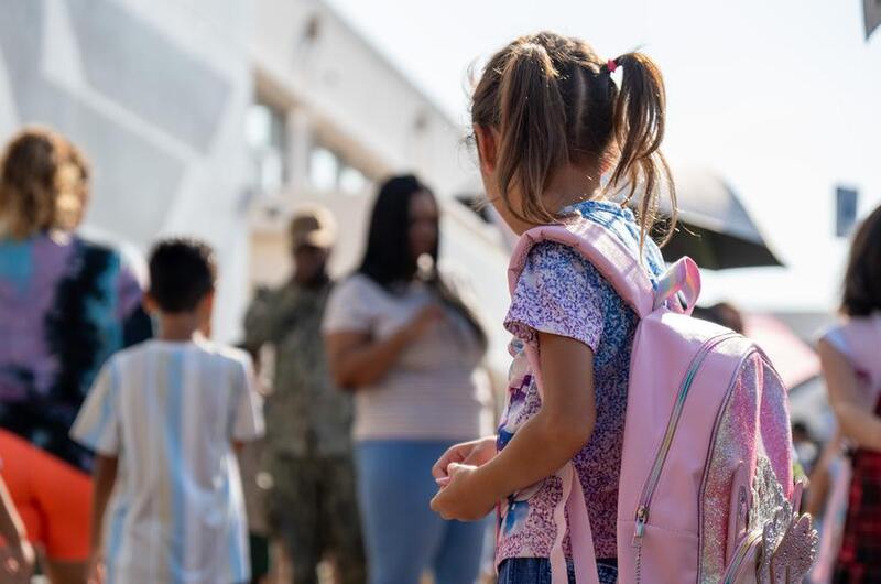A girl wearing pigtails and wearing a pink backback looks around a school yard.