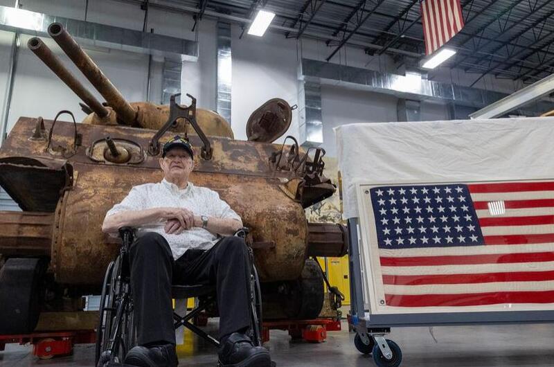A Marine veteran sits in front of a tank and beside the American flag.