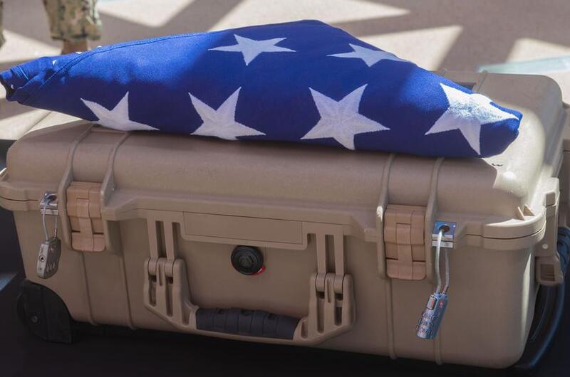 A folded American flag with white stars visible on blue fabric rests on top of a tan military-style hard case. The flag is ceremonially folded in the traditional triangular manner typically used for military honors and funeral services.
