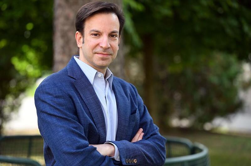 A man wearing a blue blazer and light-colored shirt stands outdoors with his arms crossed, looking at the camera. There are green trees and a blurred background behind him.