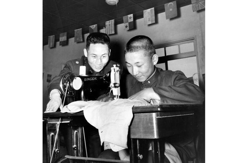 A black and white photo of a man looking on as a boy works a sewing machine on a table.