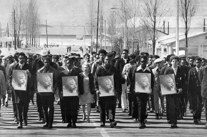 A black and white photo of people, mostly men in suits, walking while holding photos of Martin Luther King Jr.