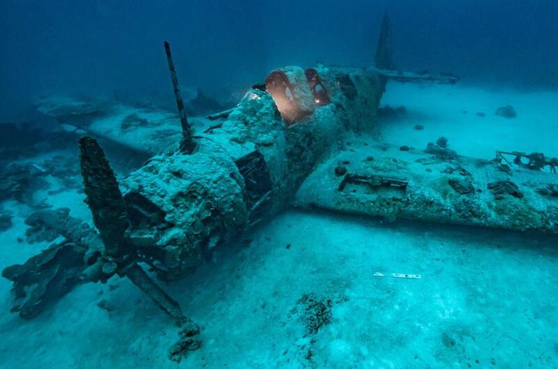 A wrecked, World War II-era military propeller plane sits on the ocean floor covered in sand and barnacles.