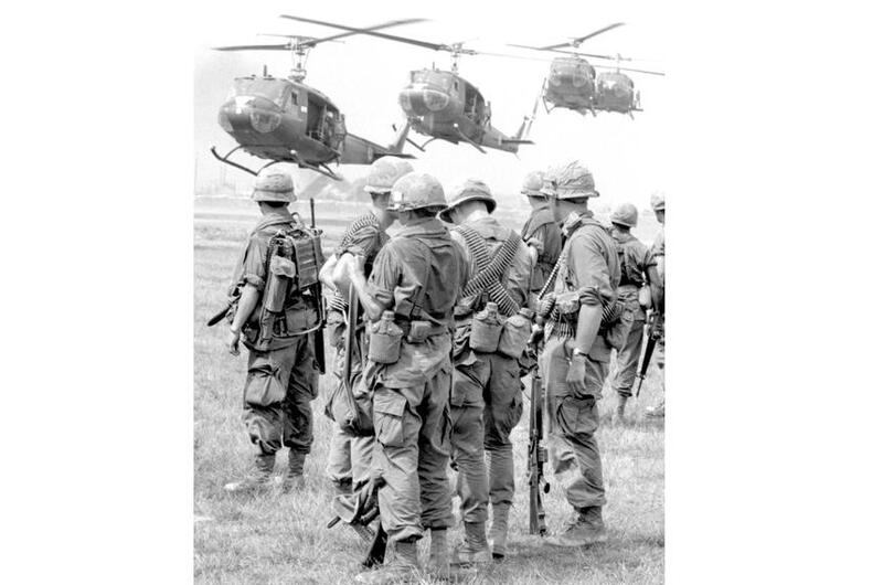 A vertical, black and white photo of service members standing as a row of helicopters flies low.