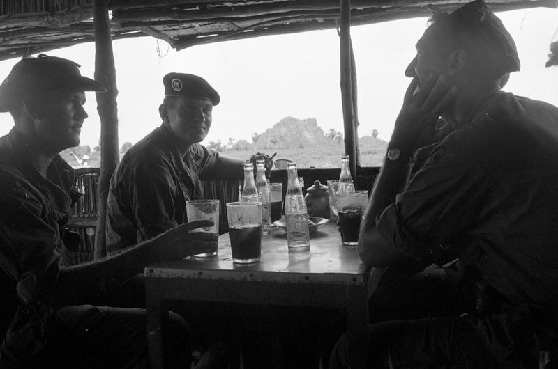 Unidentified servicemembers sit around smoking and drinking Pepsi Cola as the South Vietnamese landscape is visible behind them through the opening of the hut.