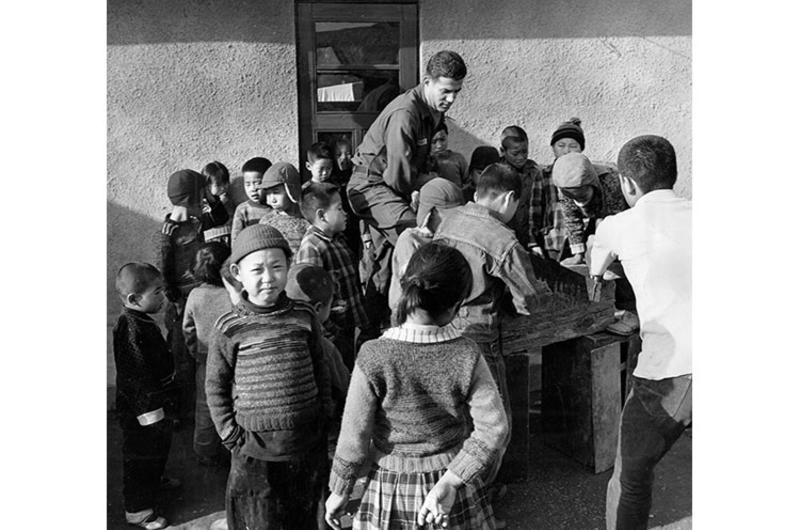 ORPHANS CLUSTER AROUND PFC SPERO METRAKOS AS HE SAWS WOOD FOR HOME'S STOVES