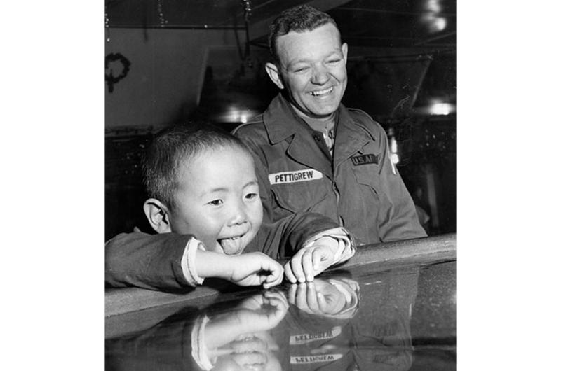 Toddler Hang Nam Chong' chins up to the bar at the Top Three Club at Ascom City as the U.S. Army Signal Depot sponsored a year-end party for orphans of the Dong Chin home which the unit supports. Delighted bystander is PFC Jimmy A. Pettigrew of the 55th Signal Co.