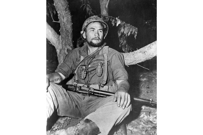 A vertical, black and white photo of a man sitting with a rifle across his lap.