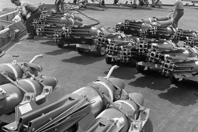 500-pound bombs are lined up on the flight deck of the USS America before being loaded onto jet planes. Visible behind the ammunition is a A-7A Corsair II (left) and a A-6A Intruder.